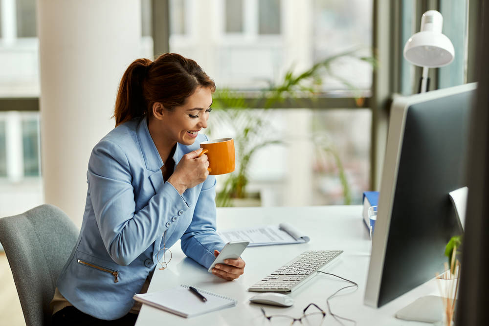 young happy businesswoman drinking coffee text messaging cell phone office